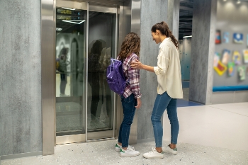 long-haired-girl-with-backpack-smiling-pleased-dark-haired-female-standing-before-closed-elevator-doors